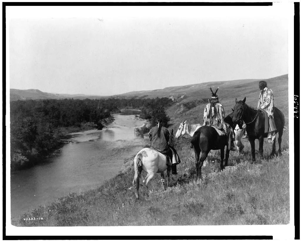 [Three Piegan Indians and four horses on hill above river], 1910